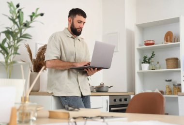 Genç sakallı adam mutfakta dizüstü bilgisayarla çalışıyor.