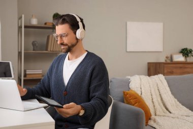 Kulaklıklı, e-okuyuculu genç adam evdeki masada dizüstü bilgisayar kullanıyor.