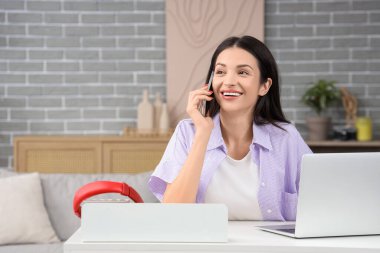 Evde dizüstü bilgisayarla cep telefonuyla konuşan güzel bir genç kadın.