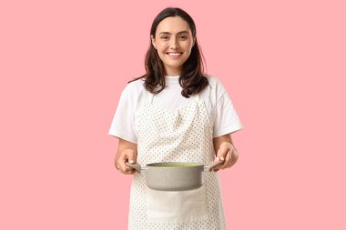 Genç ev hanımı elinde pembe arka planda lezzetli bir çorbayla yemek pişiriyor.