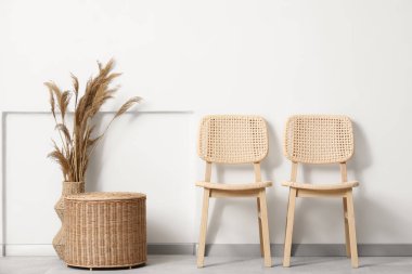 Sandalyeler, sepet ve pampalar vazoda, beyaz duvarın yanında.