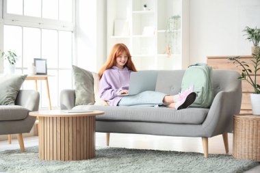 Sırt çantası ve modern dizüstü bilgisayarı olan sevimli kızıl kız evdeki kanepede uzanıyor.