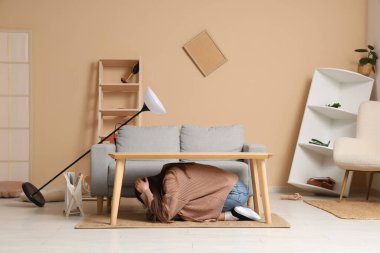 Deprem sırasında masanın altına saklanıp başını oturma odasına saklayan korkmuş genç bir kadın.