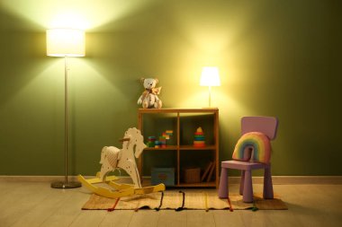 Gece çocuk odasının içinde parıldayan lambalar, sallanan at, sandalye ve yeşil duvarın yanındaki raflarda bir sürü oyuncak var.