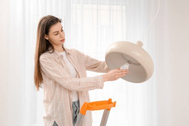 Genç bir kadın merdivende ampulü değiştiriyor. Evde şık bir lamba takıyor.