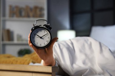 Yatak odasında, battaniyenin altında elinde retro çalar saat tutan genç bir kadın var.