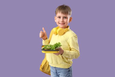 Beslenme çantasıyla leylak arka planında baş parmağını kaldıran sevimli bir okul çocuğu.