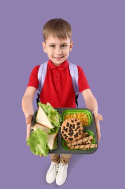 Elinde beslenme çantası, sağlıklı yiyecekler ve leylak arkaplanda tatlılar tutan sevimli bir okul çocuğu.