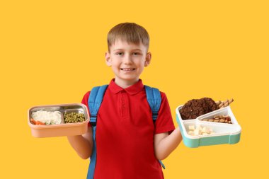 Sevimli küçük okul çocuğu beslenme çantalarını sağlıklı yiyeceklerle ve sarı arka planda tatlılar tutuyor.