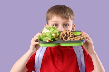 Elinde beslenme çantası, sağlıklı yiyecekler ve leylak arkaplanda tatlılar tutan sevimli bir okul çocuğu.