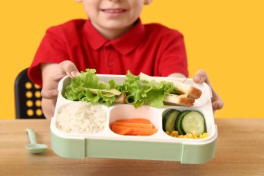 Sarı arka planda okul masasında sağlıklı yiyeceklerle beslenme çantası tutan sevimli küçük okul çocuğu.