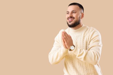 Minnettar genç adam avuçlarını birbirine bastırıyor ve bej arka planda gülümsüyor.