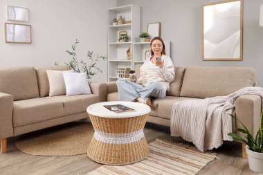 Bir fincan kahveyle güzel genç bir kadın oturma odasındaki şık gri koltukta modern cep telefonu kullanıyor.