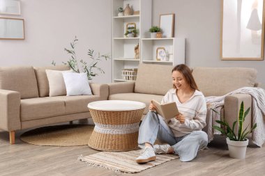 Güzel genç bayan oturma odasında şık gri kanepenin yanında kitap okuyor.