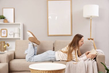 Güzel genç bayan oturma odasındaki şık gri kanepede kitap okuyor.