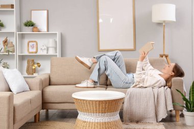 Güzel genç bayan oturma odasındaki şık gri kanepede kitap okuyor.