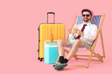 Güverte koltuğunda cep telefonu ve pembe arka planda plaj aksesuarları kullanan bir iş adamı. Yaz tatili kavramı