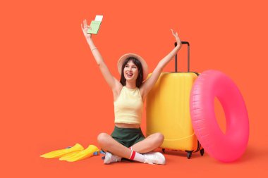 Plaj aksesuarları ve bavullu turist turuncu arka planda oturuyor.