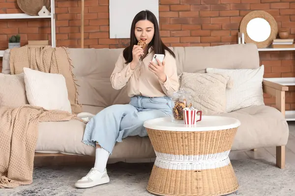 Cep telefonlu genç bir kadın evdeki kanepede kurabiye yiyor.