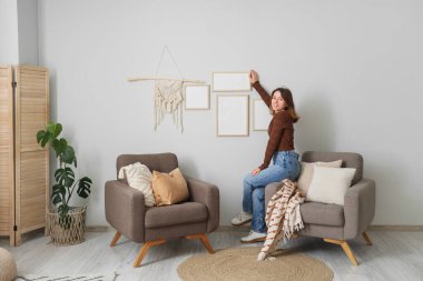 Güzel genç bir kadın oturma odasındaki beyaz duvara fotoğraf çerçevesi asıyor.