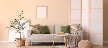 Modern oturma odasının içinde gri kanepe, kahve masası, zeytin ağacı ve bej duvarda asılı boş çerçeve.