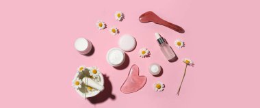 Farklı papatya çayı kozmetik ürünleri, yüz masajı aletleri ve pembe arka planda çiçekler.