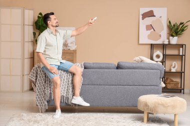 Genç bir adam oturma odasındaki bej duvarın yanındaki kanepede otururken klimayı açıyor.