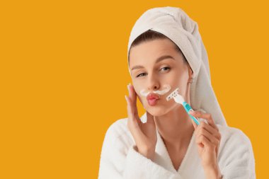 Bornozlu güzel bir kadın sarı arka planda köpük ve jiletle bıyık tıraş ediyor.