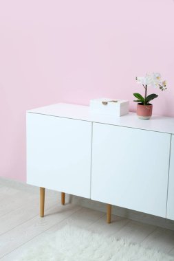 Beyaz çekmeceli, orkide çiçekli, odada pembe duvarın yanında.