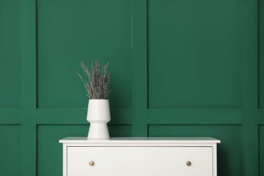 Yeşil duvarın yanındaki beyaz çekmecelerde lavantalı vazo.