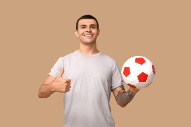 Elinde futbol topu olan yakışıklı genç adam kahverengi arka planda baş parmak hareketi yapıyor.