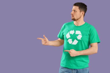 Üzerinde geri dönüşüm logosu olan tişörtlü kafası karışmış genç adam leylak arkaplanındaki bir şeyi işaret ediyor. Ekoloji kavramı