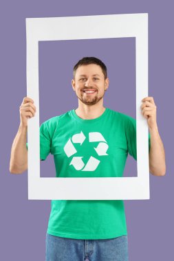 Leylak arka planında fotoğraf çerçevesi tutan geri dönüşüm amblemi olan tişörtlü mutlu genç adam. Ekoloji kavramı
