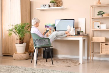 Genç bayan tasarımcı bilgisayarlı masada oturuyor ve cep telefonunu açık ofiste tutuyor.