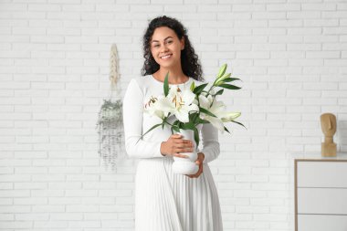 Güzel, mutlu, Afro-Amerikan bir kadın elinde bir buket beyaz zambak çiçeğiyle evinde tuğla duvarın yanında vazo tutuyor.