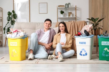 Çöpleri çöp tenekelerine dizilmiş, evde yerde oturan genç bir çift. Küresel Geri Dönüşüm Günü