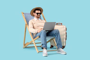 Dizüstü bilgisayarı güverte koltuğunda ve mavi arka planda bavulu olan genç bir iş adamı. Yaz tatili kavramı