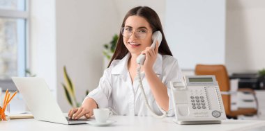 Bilgisayarlı genç sekreter masada telefonla konuşuyor.