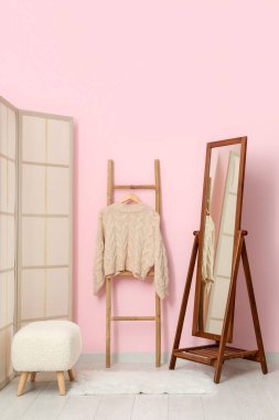 Ahşap merdivende asılı süveter, katlanan ekran, odada pembe duvarın yanında bank ve ayna.