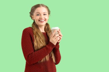 Yeşil arka planda bir fincan kahveyle sıcak süveter giyen güzel, mutlu genç bir kadın.