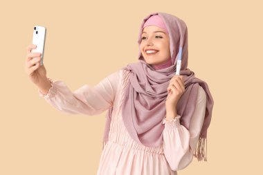 Pozitif hamilelik testi olan mutlu Müslüman genç kadın bej arka planda selfie çekiyor.