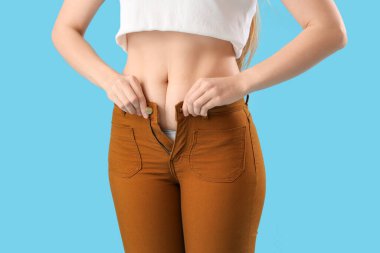 Mavi arka planda dar pantolon iliklemeye çalışan genç bir kadın, yakın plan. Kilo alma kavramı