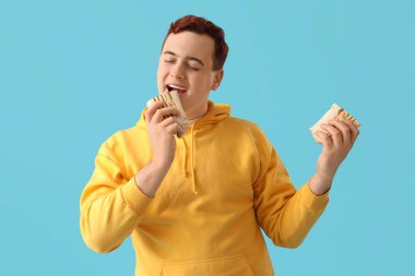 Young man eating tasty doner kebab on blue background