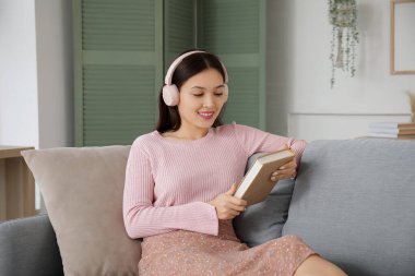 Kulaklıklı, kitapla sesli kitap dinleyen ve oturma odasında kanepede oturan güzel bir Asyalı kadın.