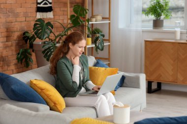 Evdeki kanepede oturan boğazı ağrıyan ve dizüstü bilgisayarı olan hasta genç bir kadın.