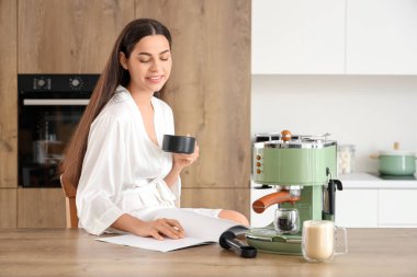 Modern kahve makinesiyle güzel, genç, mutlu bir kadın. Mutfakta kahve içip dergi okuyor.