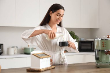Modern bir makineyle genç ve güzel bir kadın mutfakta bir fincan taze kahveye süt dolduruyor.