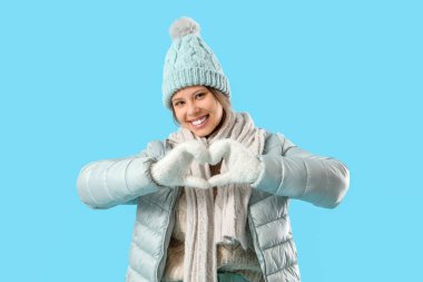 Kışın giyinmiş genç bir kadın mavi arka planda kalp hareketi yapıyor.