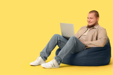 Laptoplu genç adam sarı arka planda fasulye koltukta oturuyor.