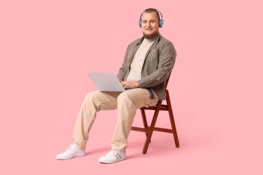 Kulaklıklı, dizüstü bilgisayarlı, pembe arka planda sandalyede oturan genç adam.